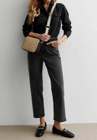 Donna con camicia nera con bottoni, jeans neri a vita alta, mocassini neri e borsa a tracolla con texture intrecciata e manico largo.