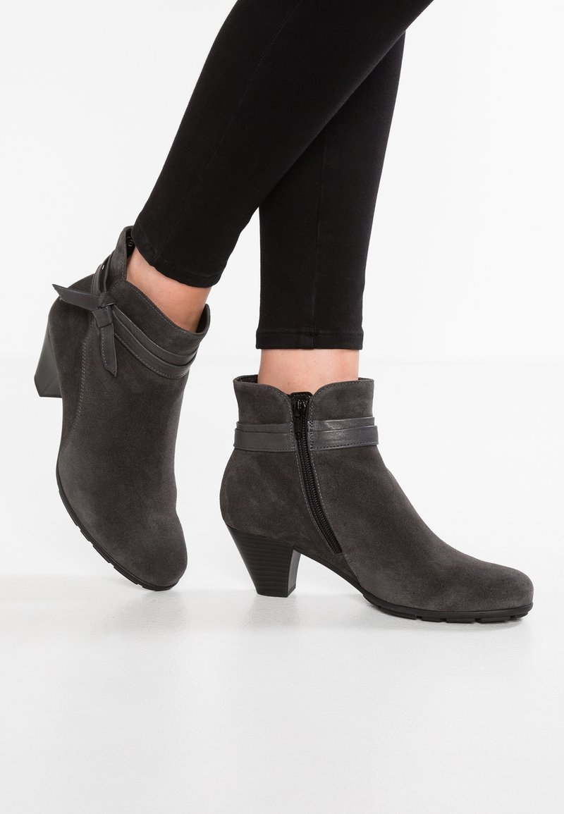Femme portant un pantalon noir slim et des bottines en daim gris foncé avec talons blocs, fermeture éclair latérale et détail de sangle en cuir.