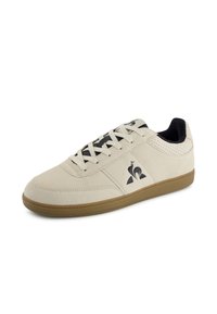 Sneaker in suede beige con logo nero, punta tonda e dettagli perforati sui lati. Presenta lacci grigi e suola in gomma color gomma.