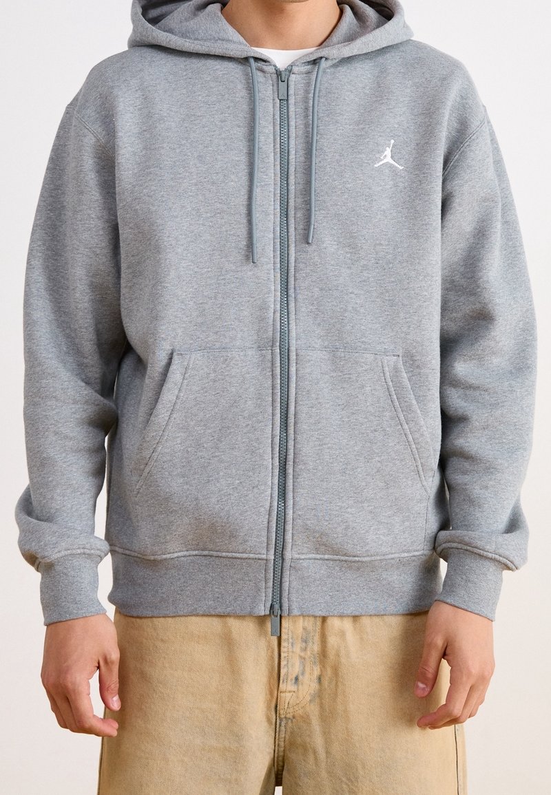 Homme portant un sweat à capuche gris clair zippé avec poches avant et un petit logo blanc sur la poitrine gauche, associé à un pantalon beige.