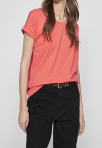 Korálové tričko s krátkým rukávem a ohrnutými rukávy, vyrobené z měkké bavlny. V kombinaci s černými kalhotami a hnědým páskem s zlatou sponou.