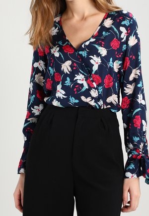 Mujer vistiendo una blusa cruzada floral azul marino con flores rojas, blancas y verde azulado, combinada con pantalones negros de tiro alto.