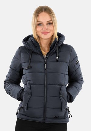 Femme portant une veste à capuche matelassée bleu marine avec des poches zippées et à pression, souriant avec les mains dans les poches contre un fond uni.