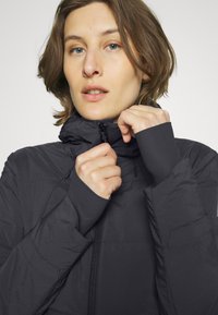 Schwarze isolierte Jacke mit hohem Kragen, Reißverschluss vorne und Daumenlöchern an den Ärmelbündchen. Glatte, matte Textur mit dezenten Nähten.