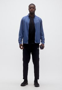 Νεαρός άνδρας που φοράει μπλε ζακέτα με φερμουάρ, μαύρο πουκάμισο, μαύρο παντελόνι και μαύρα αθλητικά παπούτσια, κρατώντας ένα μπουκάλι νερό στο δεξί χέρι.