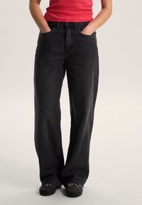 Černé džínové kalhoty s širokými nohavicemi a vysokým pasem, předními kapsami a jemnými švy, v kombinaci s přiléhavým červeným tílkem.