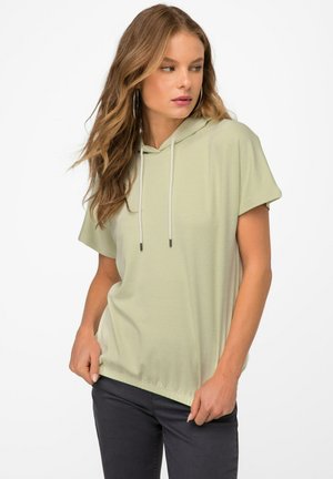 Hoodie vert clair à manches courtes avec capuche et cordons de serrage, présentant un tissu texturé et une coupe décontractée, associé à un pantalon sombre.