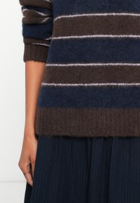 Kar csíkos barna, tengerészkék és levendula színű pulóver mellett, bordázott szegéllyel, ami egy sötét pliszírozott szoknya fölött van, világos háttér előtt.