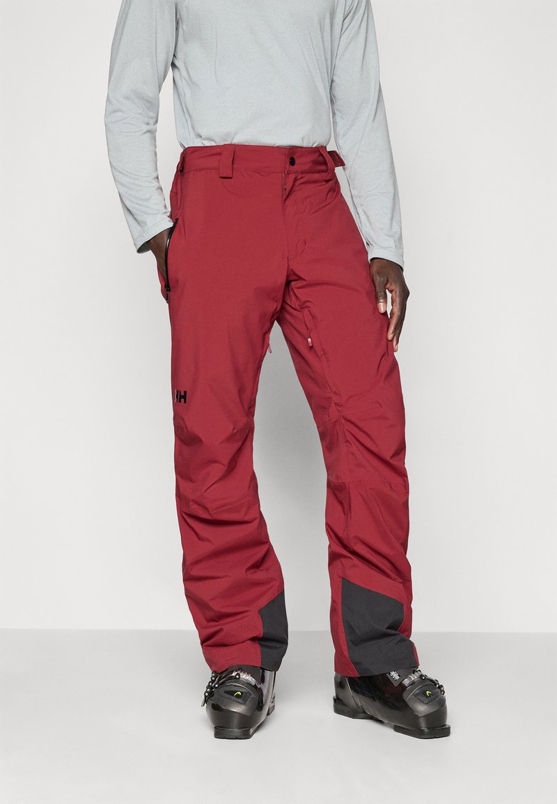Homme portant un pantalon de ski rouge et des bottes de ski noires, se tenant avec une main dans la poche contre un fond clair et sobre.