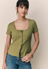 Ribbad grön kortärmad topp med fyrkantig necklinje och knappad front. Matchad med blå denimjeans. Slät textur genom hela plagget.