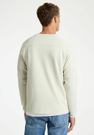 Man die een gebroken witte lange mouwen shirt en blauwe spijkerbroek draagt, gezien van achteren tegen een effen witte achtergrond.