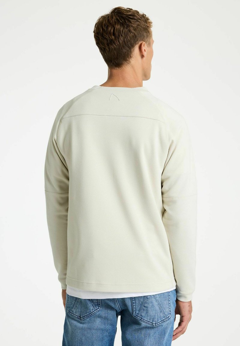 Moški v dolgo rokavski srajci barve slonovine in modrih kavbojkah, prikazan od zadaj na enobarvni belem ozadju.