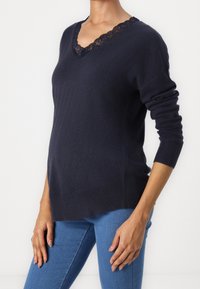 Kvinna iförd en mörk marinblå V-ringad tröja med spetsdetaljer, kombinerad med figursydda blå jeans, stående mot en enfärgad vit bakgrund.