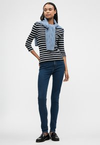 Ριγέ μπλούζα με μακριά μανίκια σε ναυτικό μπλε και λευκό, συνδυασμένη με σκούρα μπλε κολάν παντελόνι. Μια ανοιχτό μπλε πλέκη κασκόλ κρέμεται χαλαρά γύρω από τον λαιμό. Μαύρα παπούτσια.