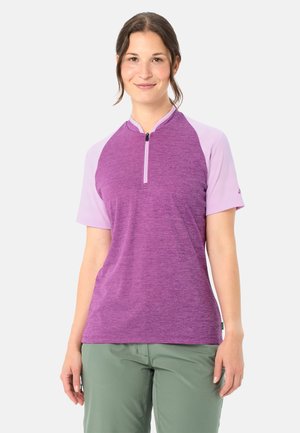 Junge Frau steht lächelnd da, trägt ein lila kurzärmeliges Zip-up-Shirt und grüne Hosen vor einem schlichten hellen Hintergrund.