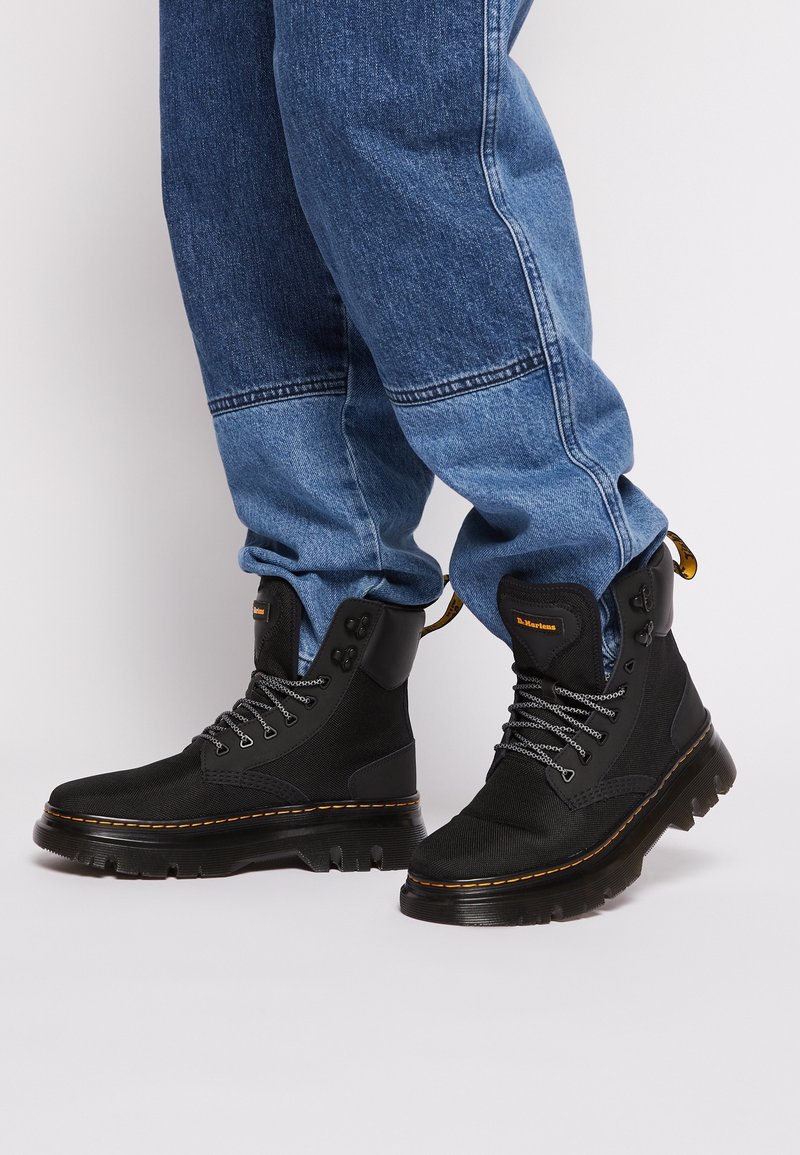 Personne portant des bottes noires à lacets avec semelles épaisses et un jean bleu en denim sur un fond clair uni.