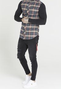 Camicia a quadri con maniche corte nere, abbinata a jeans neri strappati e sneakers bianche. Presenta un'etichetta appesa ai jeans.