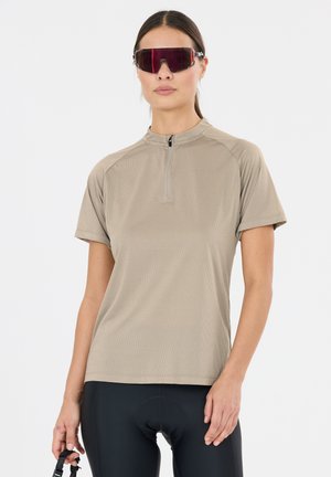 Vrouw draagt een beige wielertrui met korte mouwen en zwarte gewatteerde fietsbroek, houdt een zonnebril in één hand tegen een witte achtergrond.