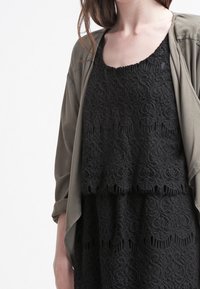 Svart spetsblus med lagerdesign, med blommönster och vågiga kanter, kombinerad med en olivgrön, lätt jacka.