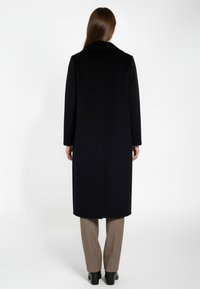 Kvinna som står med ryggen mot kameran, iklädd en lång svart kappa, beige byxor och svarta klackskor framför en enkel vit bakgrund.