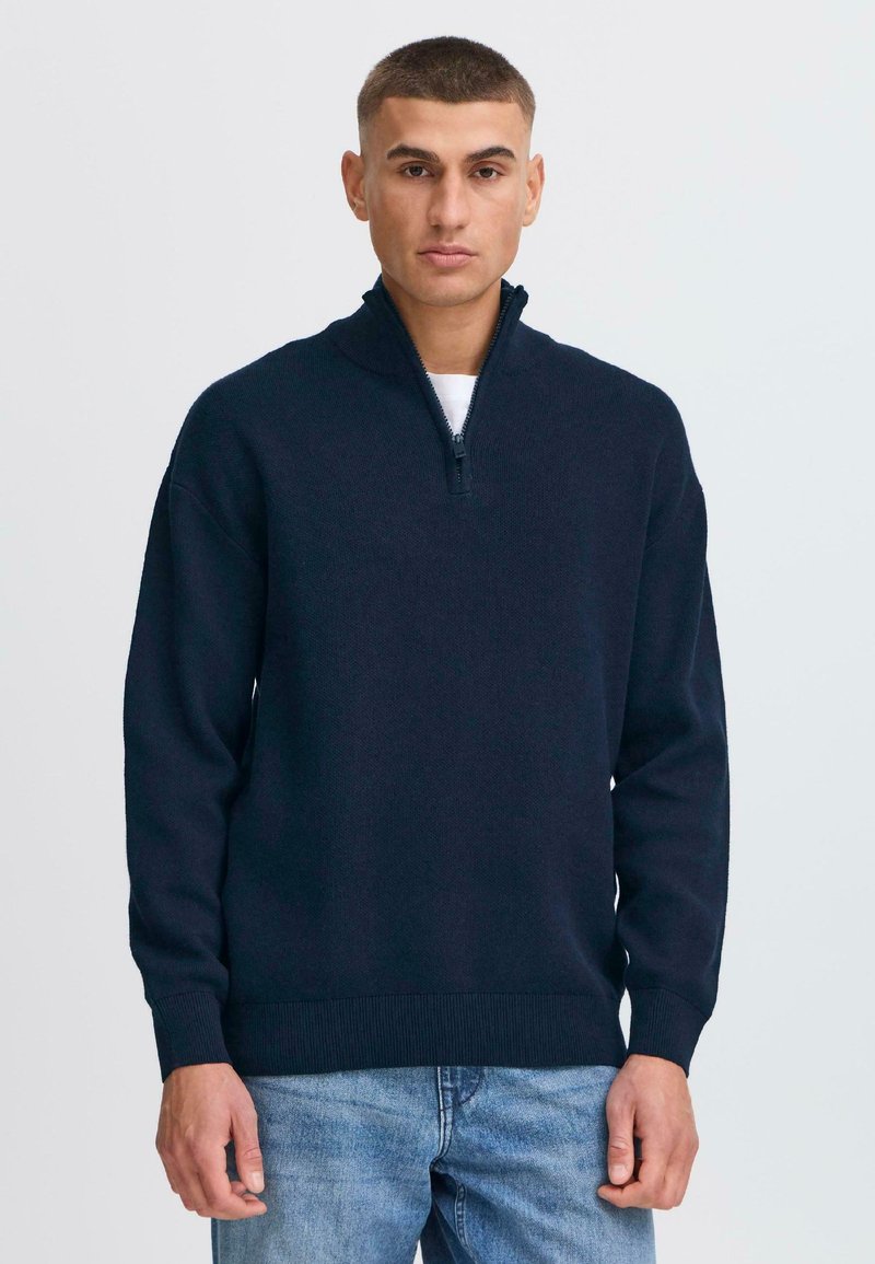Pullover in maglia blu navy scuro con colletto a zip quart, polsini e orlo a coste, indossato sopra una maglietta bianca, abbinato a jeans azzurri chiari.
