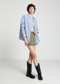 Chemise à rayures bleues et blanches boutonnée, portée sur un haut gris, mini jupe verte et bottes noires montantes. Tissu lisse avec des couleurs contrastées.