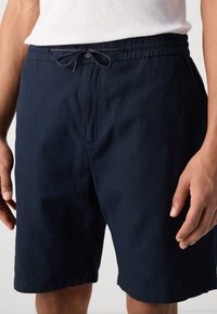 Pantaloni scurți de culoare albastru marin, realizați dintr-un material moale și ușor. Au un brâu elastic cu șnur ajustabil și buzunare laterale.