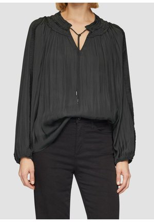 Femme portant un chemisier noir ample à manches longues avec des poignets et un col froncés, associé à un pantalon noir sur un fond uni.