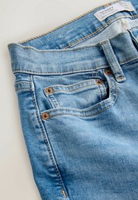 Jean skinny en denim bleu clair montrant la poche avant, la boucle de ceinture et une étiquette visible avec les détails de taille.