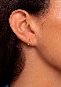 Goldene Creole mit kleinen, kugelförmigen Akzenten, die am Ohr eines Modells vor einem glatten Hauthintergrund präsentiert wird.