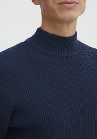 Temno modra pletena majica z visokim rebrastim ovratnikom in teksturiranim vafljem. Poudarjen bližnji pogled, ki izpostavlja materiale in oblikovalske podrobnosti.