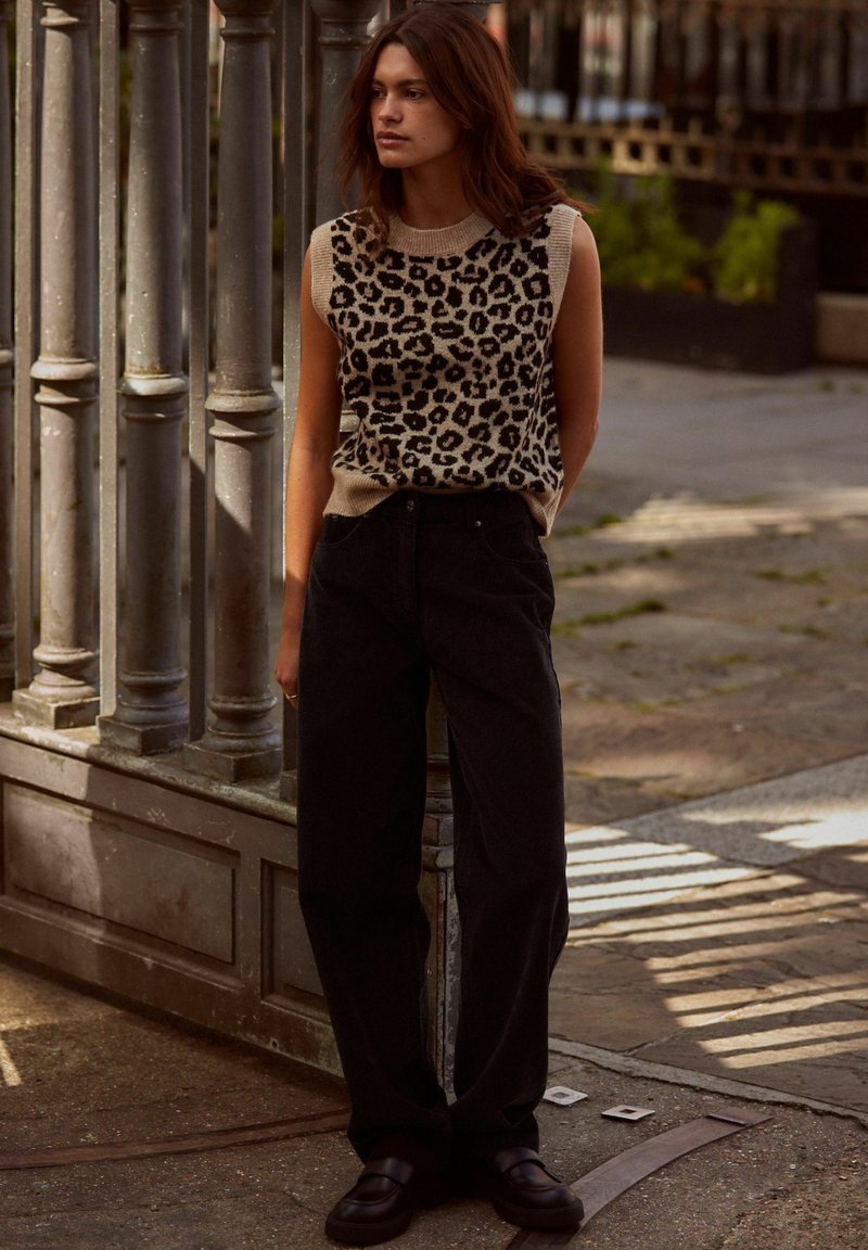 Pull en tricot avec imprimé léopard beige et noir, associé à un pantalon noir taille haute et des chaussures noires imposantes. Se tenant contre une rambarde.