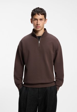 Jeune homme portant un pull marron zippé et un pantalon foncé, debout les mains dans les poches devant un fond blanc uni.