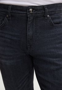 Jean en denim foncé avec une texture lisse, présentant une fermeture à bouton à l'avant, des passants de ceinture et un léger décoloration sur le tissu.