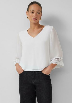 Frau trägt eine weiße V-Ausschnitt-Bluse mit 3/4-Ärmeln mit Spitzenbesatz und schwarze Hose, steht vor einem hellgrauen Hintergrund.