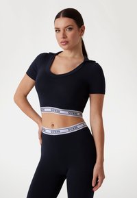 Top corto negro con mangas cortas y escote redondeado. Presenta una cinturilla de logo de marca en contraste blanco. Textura suave y diseño ajustado.