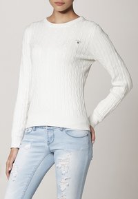 Witte kabelgebreide trui met een ronde hals en geribbelde randen, met een klein logo op de voorkant, gecombineerd met lichtblauwe distressed jeans.