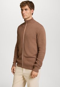Brun strik-zippesweater med stående krave, der har et struktureret mønster og en orange accent langs lynlåsen. Båret over lyse bukser.