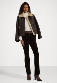 Veste en cuir marron avec un col en fourrure, deux poches avant et une coupe carrée. Stylée avec un pull col roulé beige et un pantalon en velours côtelé marron foncé.