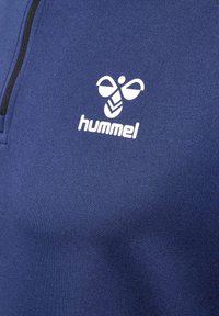 Navyblaues Poloshirt mit halbem Reißverschluss und einem weißen Logo auf der Brust. Der Stoff scheint glatt und atmungsaktiv zu sein.
