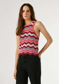 Νεαρή γυναίκα με μακριά καστανά μαλλιά, φορεμένη με αμάνικη μπλούζα με πολύχρωμο ζιγκ-ζαγκ μοτίβο και ψηλόμεσο μαύρο παντελόνι, στέκεται μπροστά σε λευκό φόντο.