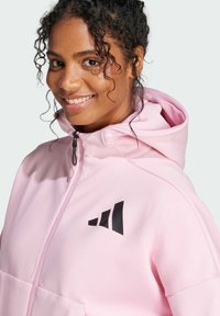 Giacca con cappuccio rosa in tessuto texturizzato, con un logo nero nella parte superiore sinistra. Dettagli con zip e tasche frontali sono visibili.
