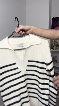 Sweater branco com riscas horizontais pretas, textura canelada e colarinho. Apresenta um design de decote em V e está pendurado num cabide preto.