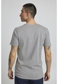 Camiseta de algodón gris con cuello redondo y mangas cortas, con un ajuste relajado. Tatuajes visibles en los brazos, sin detalles de diseño prominentes.
