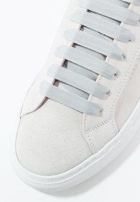 Baskets en daim gris clair avec larges lacets plats gris et semelle en caoutchouc blanche texturée, présentées en gros plan de la zone des orteils.