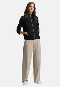 Woman standing with hands in pockets wears black zip-up jacket, beige wide-leg pants, and white sneakers on a plain background.