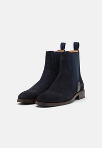 Paire de bottines Chelsea en daim bleu marine foncé avec talons bas marron et panneaux latéraux élastiques noirs sur fond blanc.