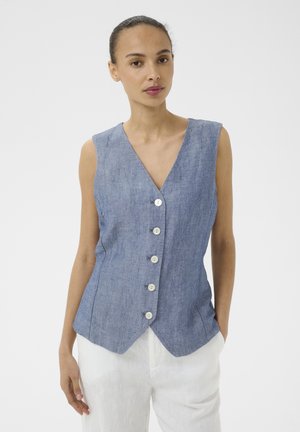 Femme portant un gilet bleu sans manches à boutons avec un col en V et un pantalon blanc, debout devant un fond blanc uni.