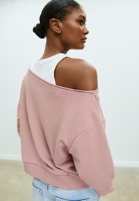 Felpa rosa oversize con scollatura ampia e spalle cadenti. Abbinata a una canotta bianca. Tessuto morbido, design casual.