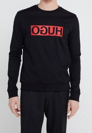 Mężczyzna ubrany w czarną bluzę z długim rękawem z odwróconym czerwono-czarnym logo "HUGO" na klatce piersiowej, w czarnych spodniach, stojący na jednolitym tle.